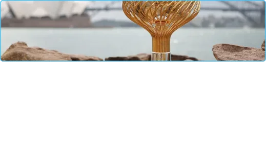 Sculpture en spirale dorée sur un premier plan rocheux avec une vue floue de l'Opéra de Sydney et du Harbour Bridge en arrière-plan. La scène est sereine et artistique.